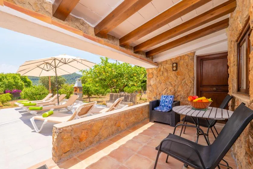 A covered stone patio with outdoor seating overlooks a sunny poolside garden.