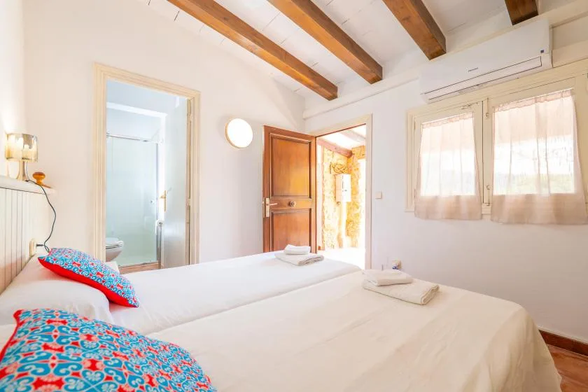 A bright bedroom with two beds, colorful pillows, and exposed wooden ceiling beams.