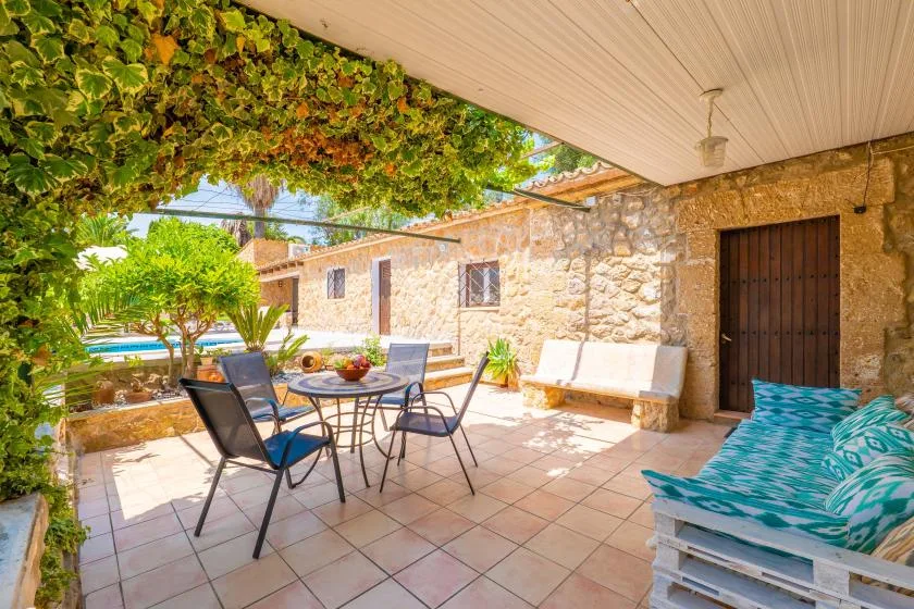 A covered stone patio has an outdoor dining table and seating beside a blue pool.