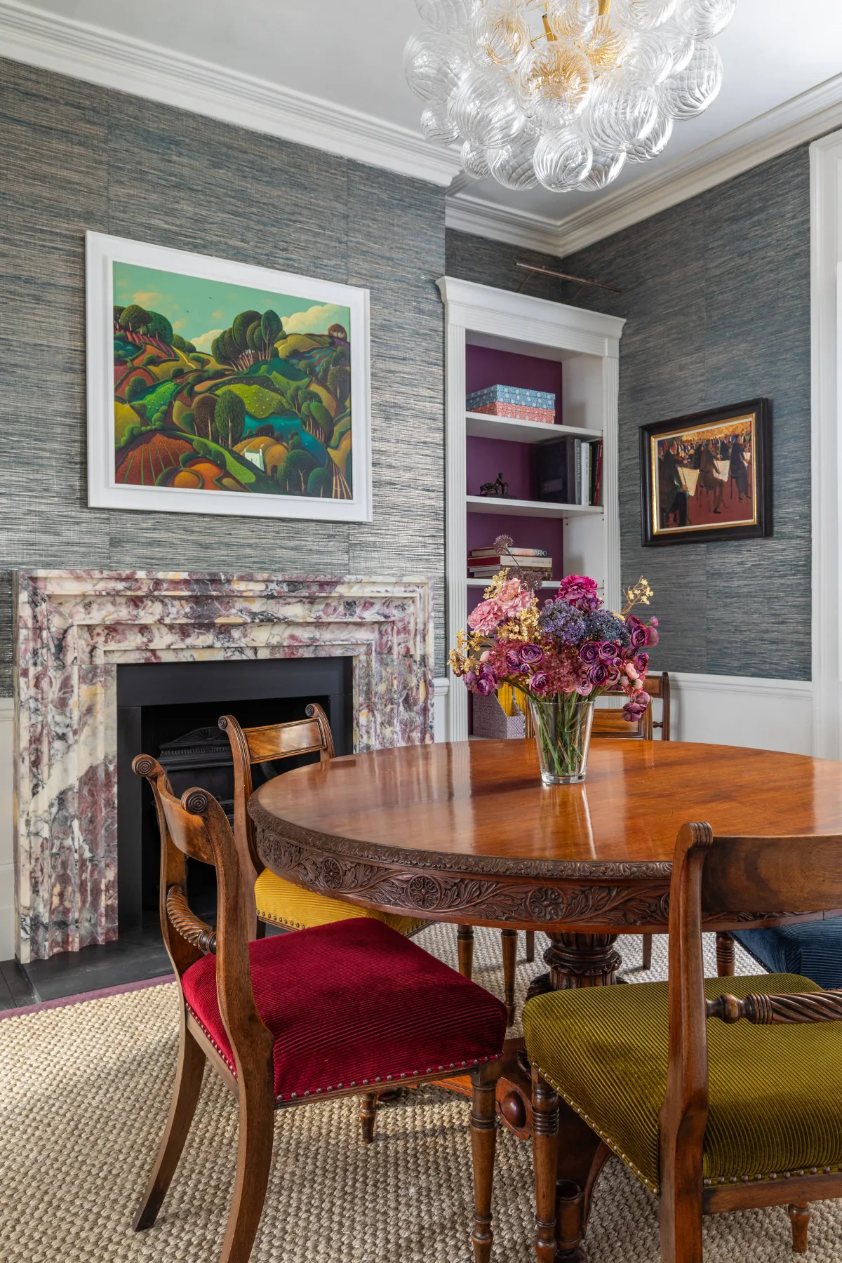 Comedor con una mesa de madera tallada, sillas coloridas y una chimenea.
