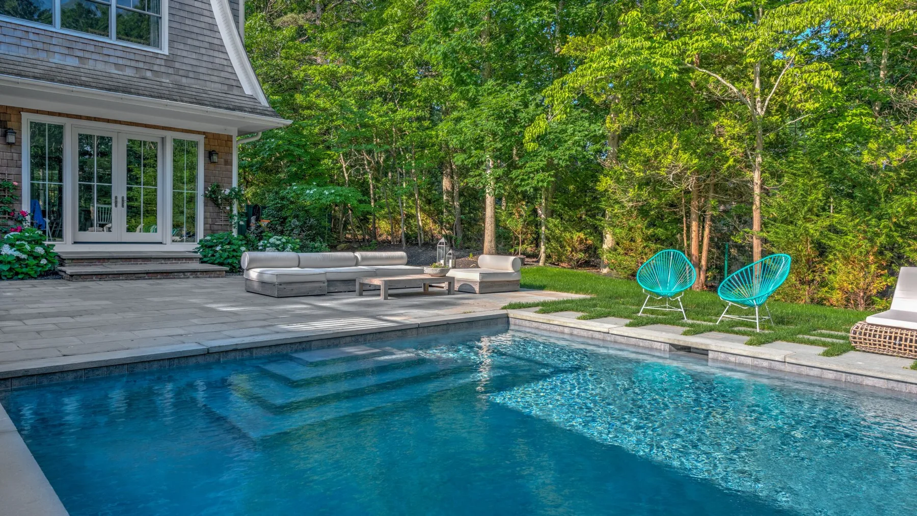 Una piscina junto a un patio, con asientos y árboles verdes al fondo.