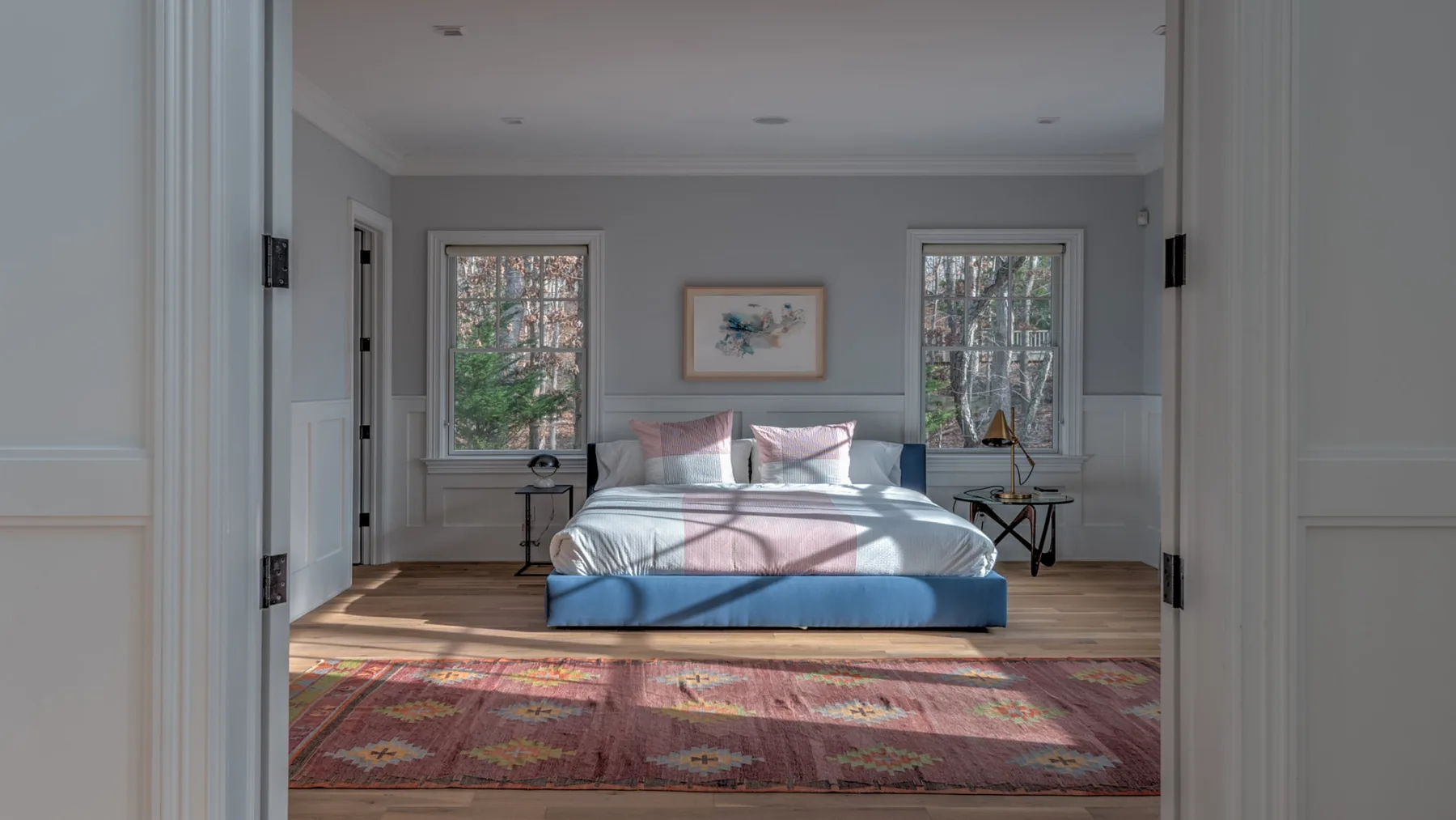 Un dormitorio con una cama bajo un cuadro enmarcado y dos ventanas.