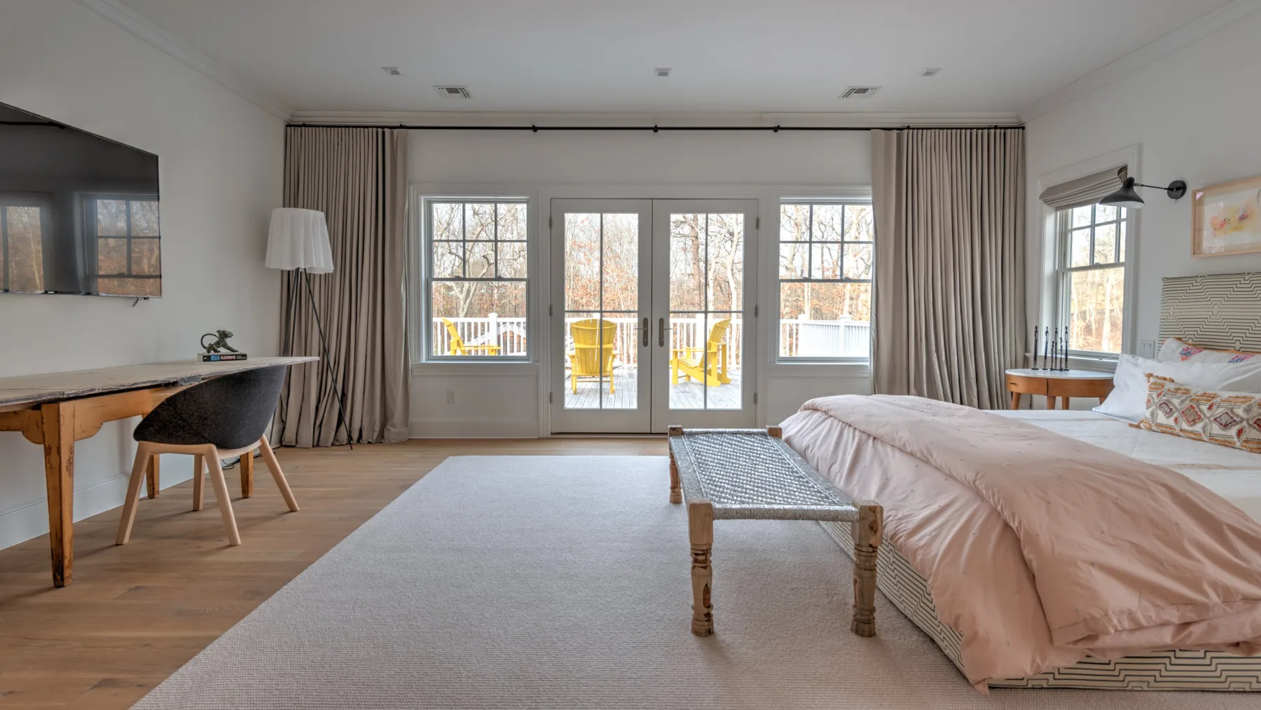 Un dormitorio luminoso con cama, escritorio y grandes ventanas que dan a un balcón.