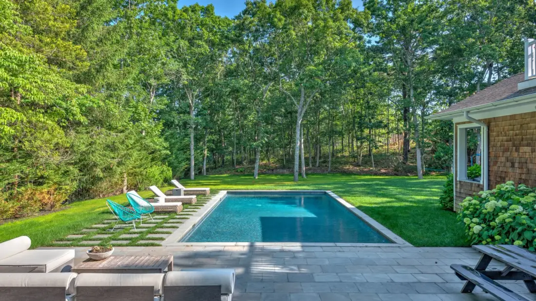 Una piscina al aire libre está junto a un patio pavimentado y vegetación arbolada.