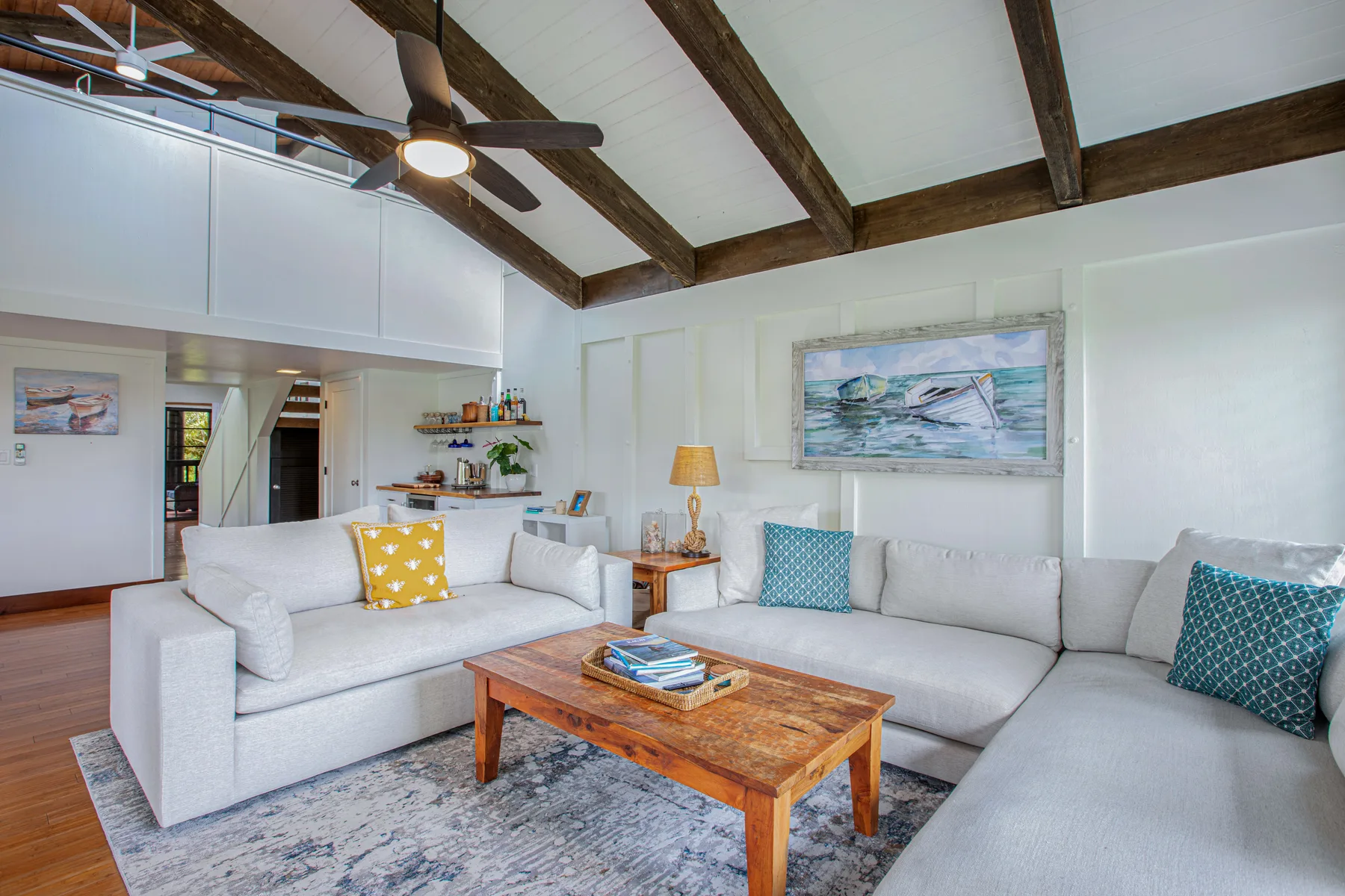 A bright living room with light-colored seating and a wooden coffee table beneath exposed ceiling beams.