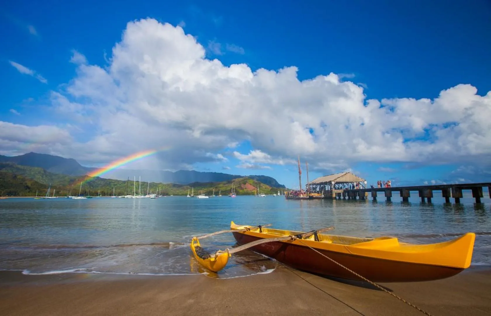 A sandy shoreline with colorful boats, calm water, and a rainbow over distant hills.