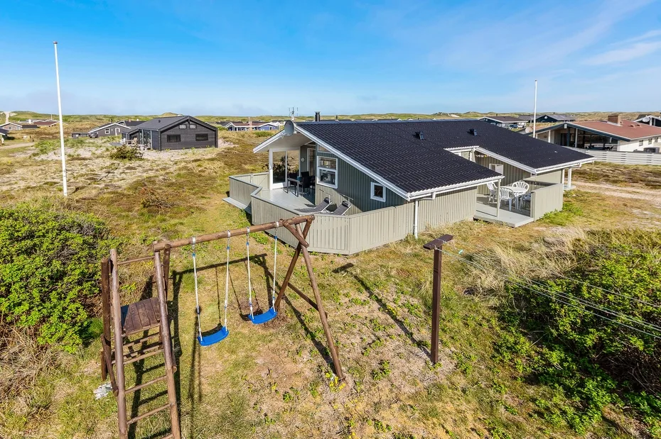 Una casa costera independiente con terraza y columpios en un paisaje arenoso.
