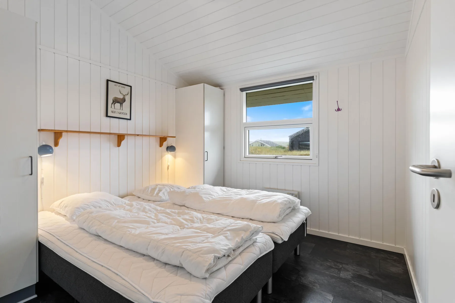 Un dormitorio luminoso con ropa de cama blanca, un estante de pared y una ventana.
