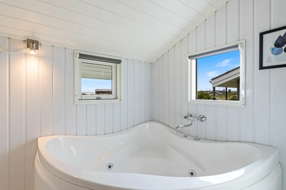 Un baño luminoso con una amplia bañera de esquina bajo dos ventanas.