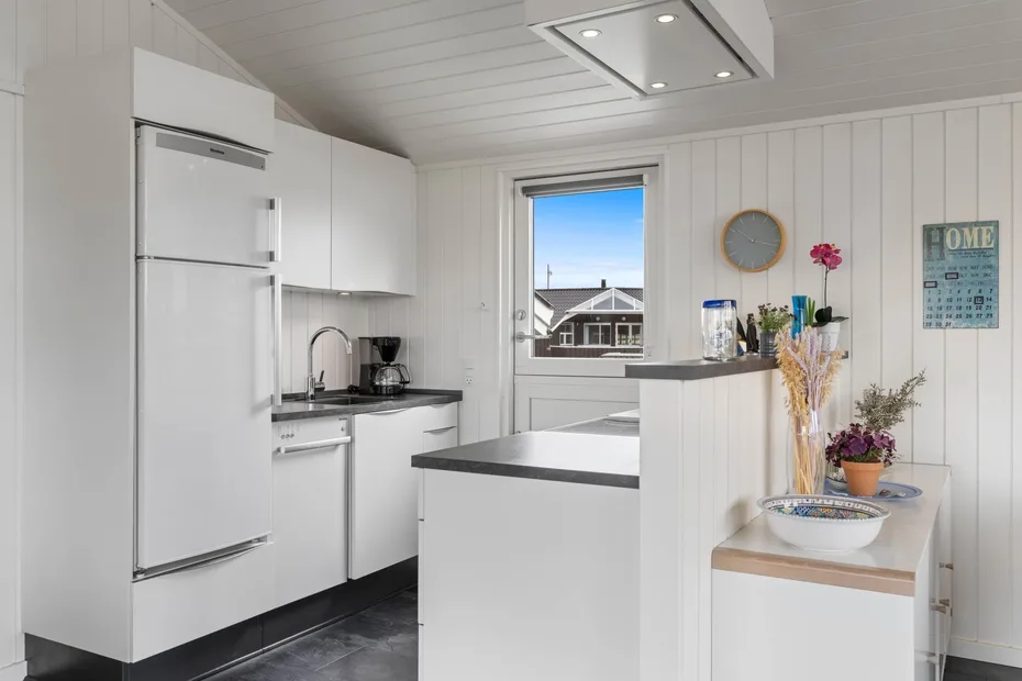 Una cocina blanca y luminosa con una isla central y una ventana con vista a los tejados.