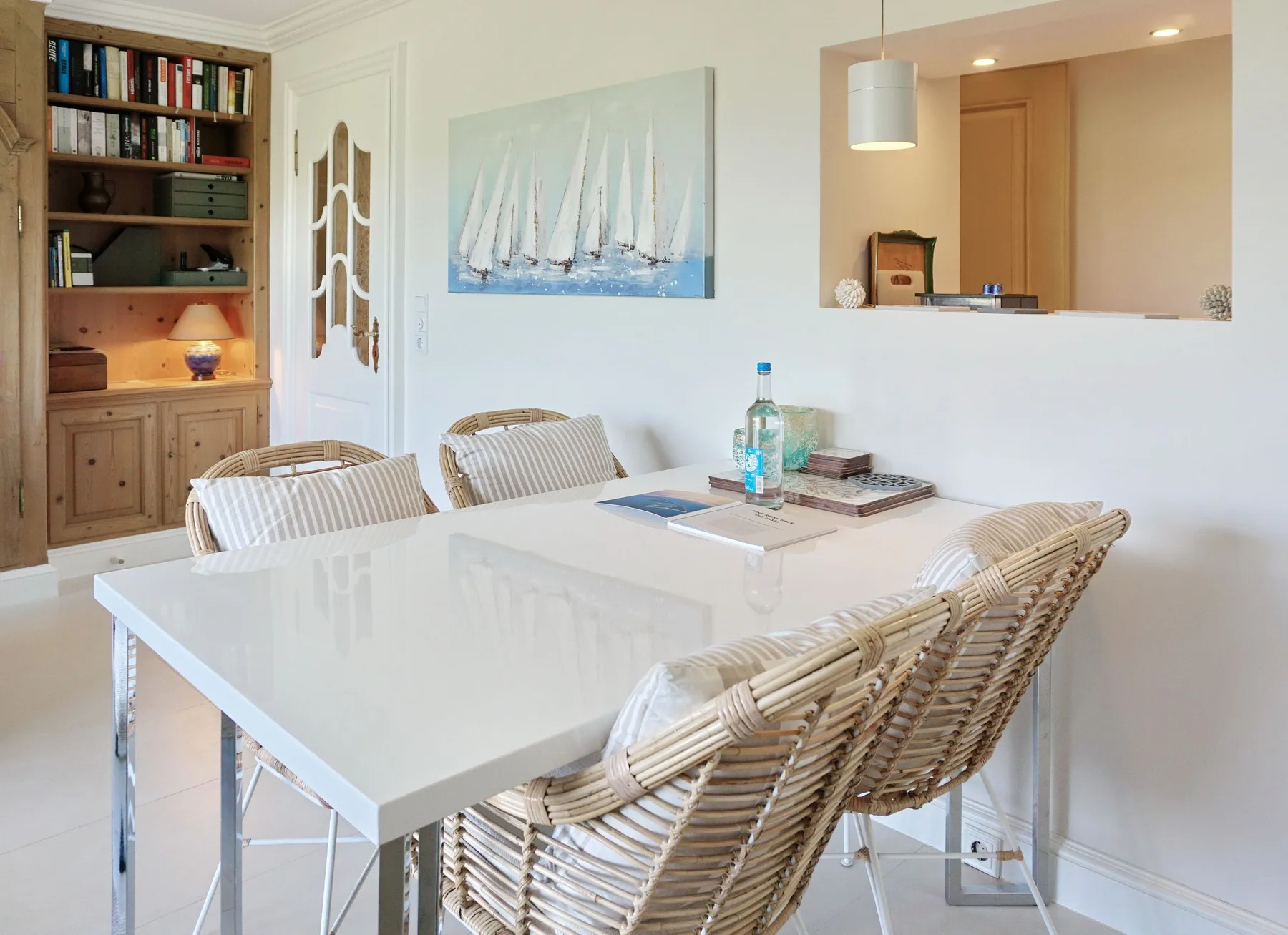 Un comedor luminoso con una mesa blanca, asientos tejidos y un cuadro de veleros.