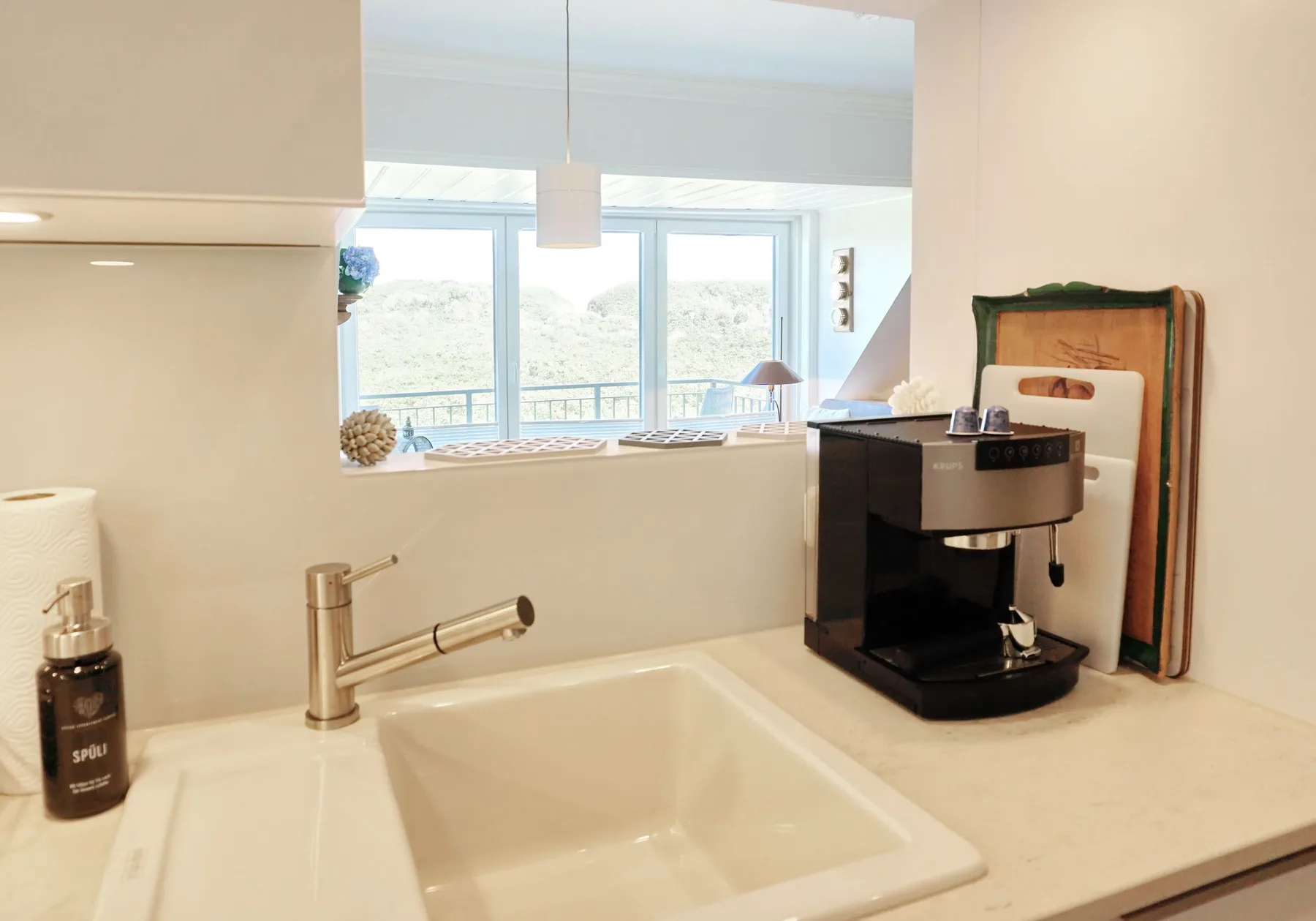 Un lavabo y una encimera de baño están junto a una cafetera, con una ventana luminosa al fondo.
