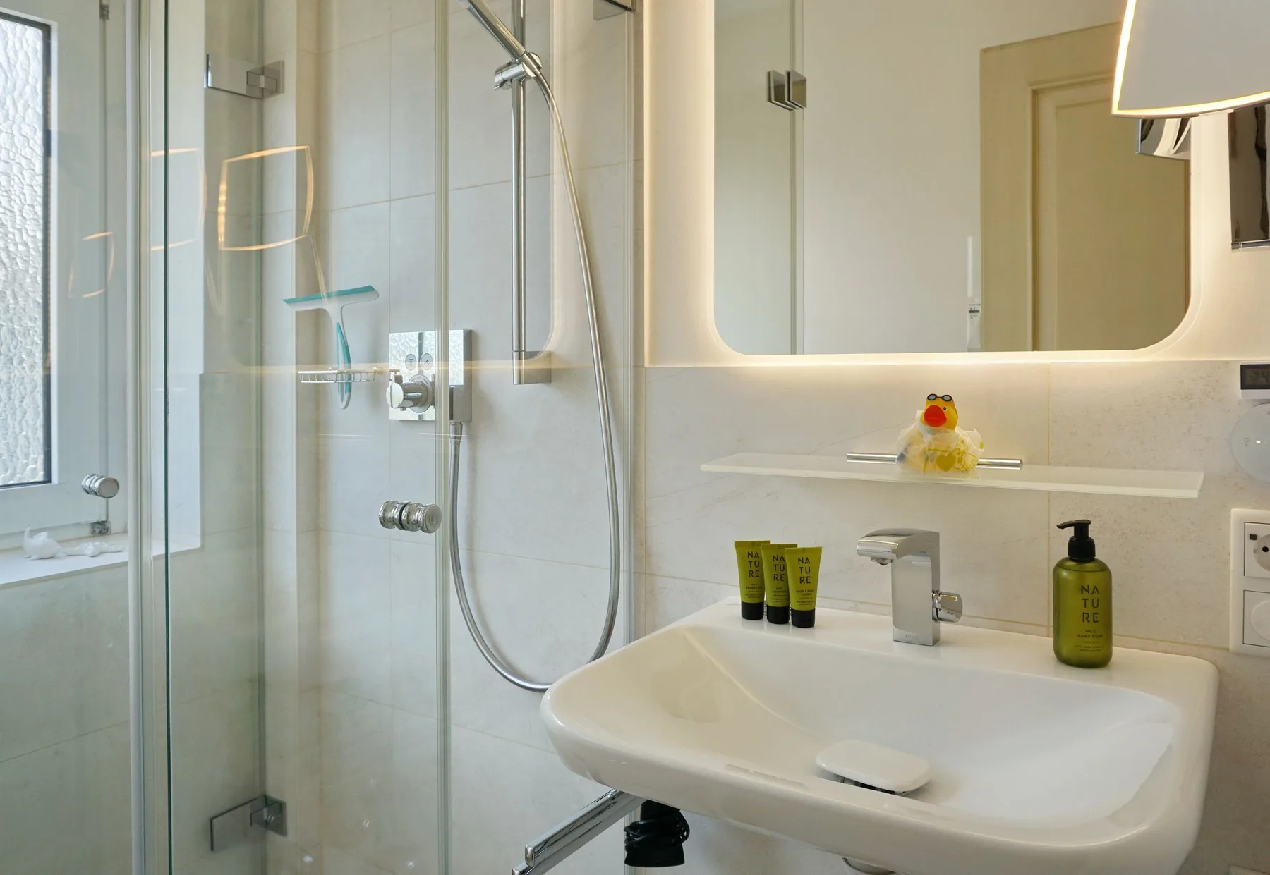 Un baño luminoso con una ducha de vidrio y un lavabo bajo un espejo iluminado.
