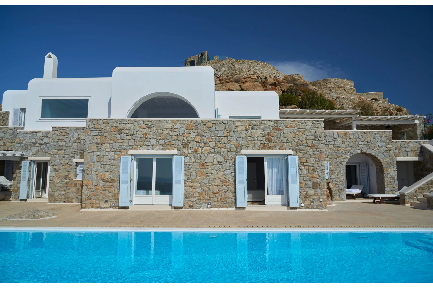 A stone villa with white upper walls overlooks a bright blue swimming pool.