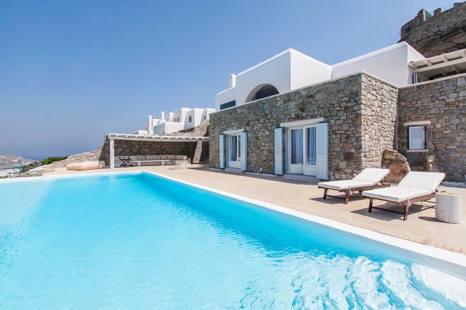 A swimming pool beside a stone-faced villa with white lounge seating.