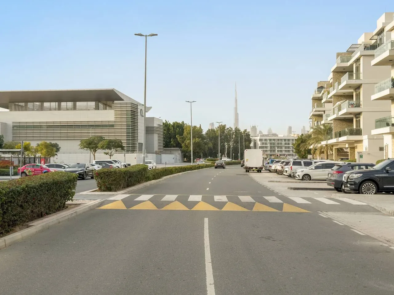 Una calle entre edificios modernos, con coches aparcados y una torre a lo lejos.