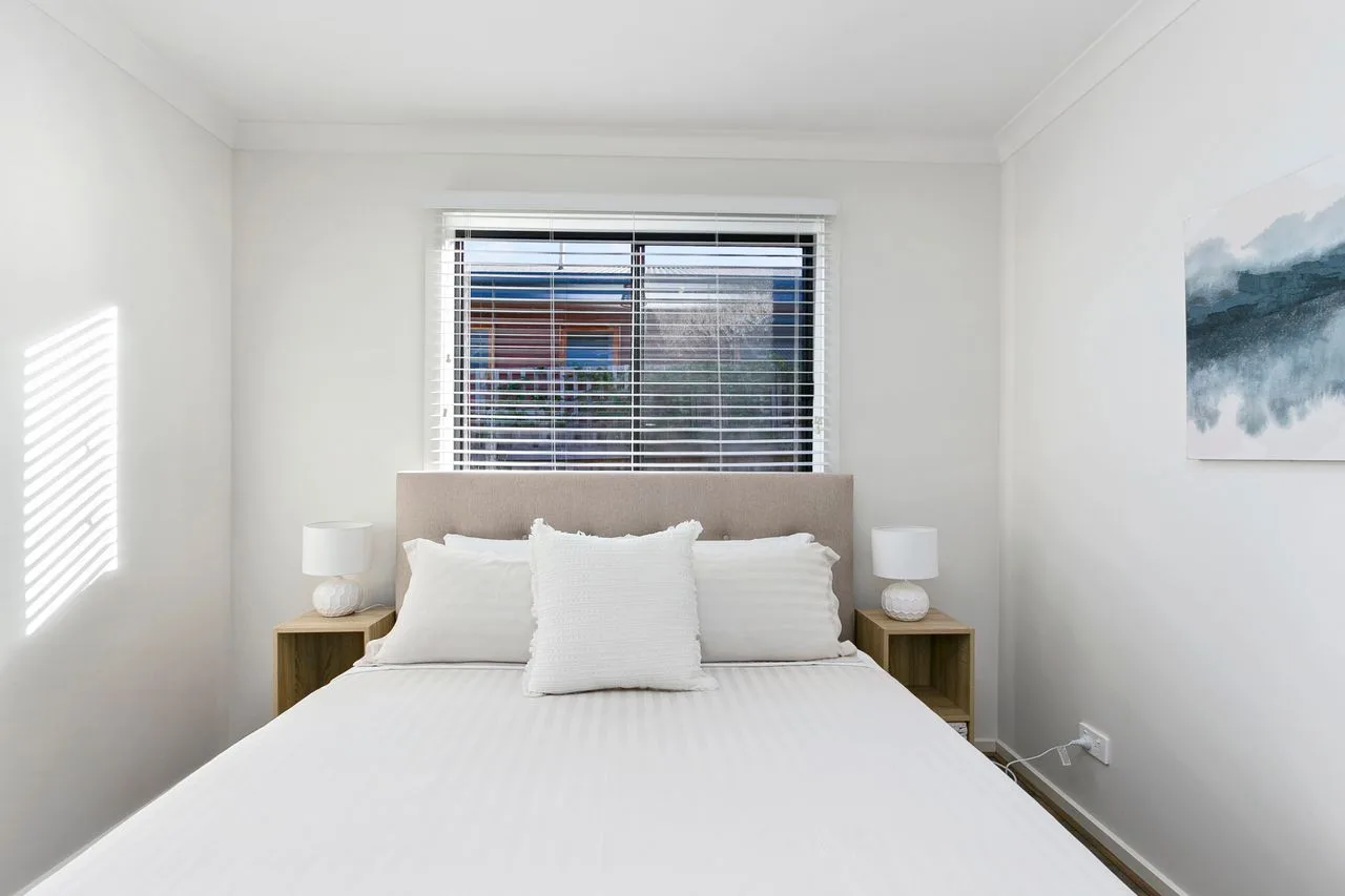 A bright bedroom with a neatly made bed beneath a window with blinds.