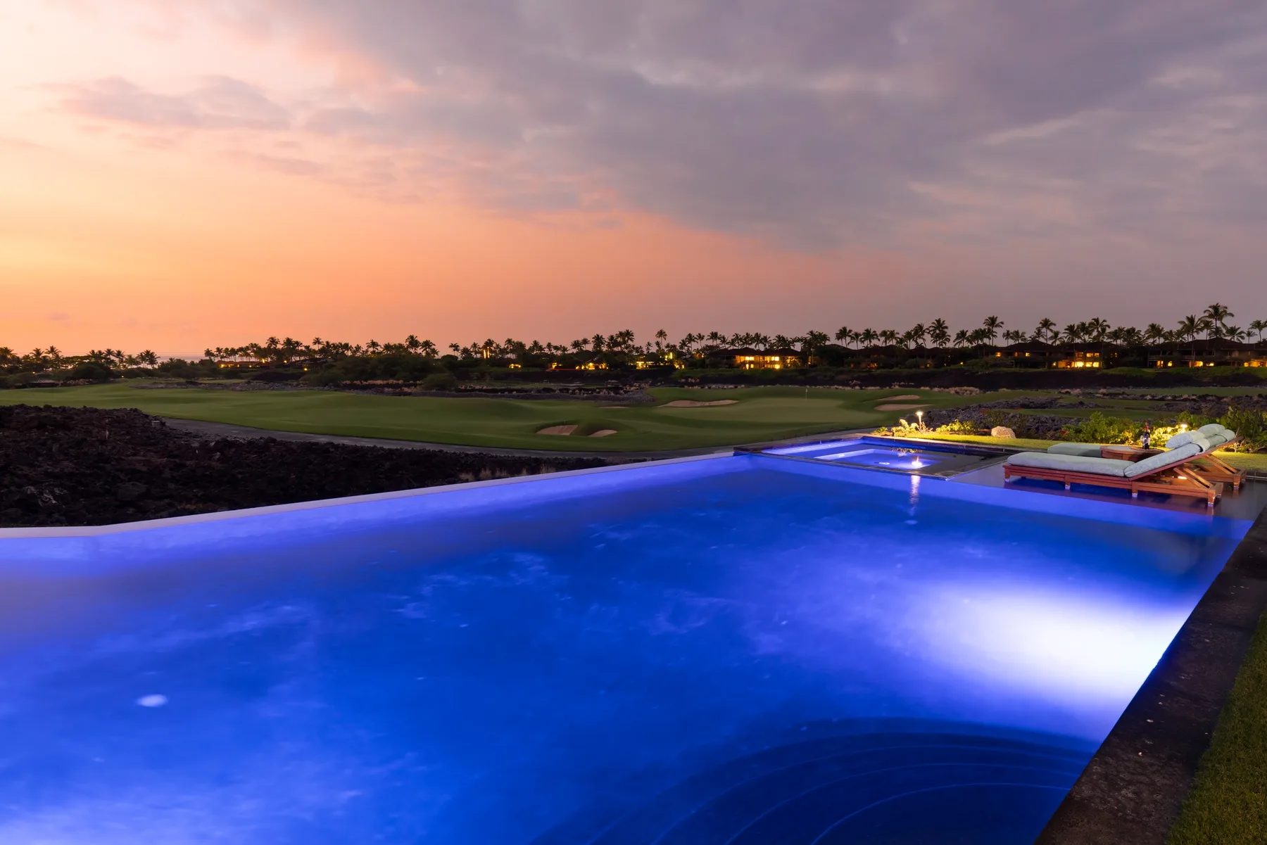 An illuminated swimming pool overlooks a golf course at sunset.