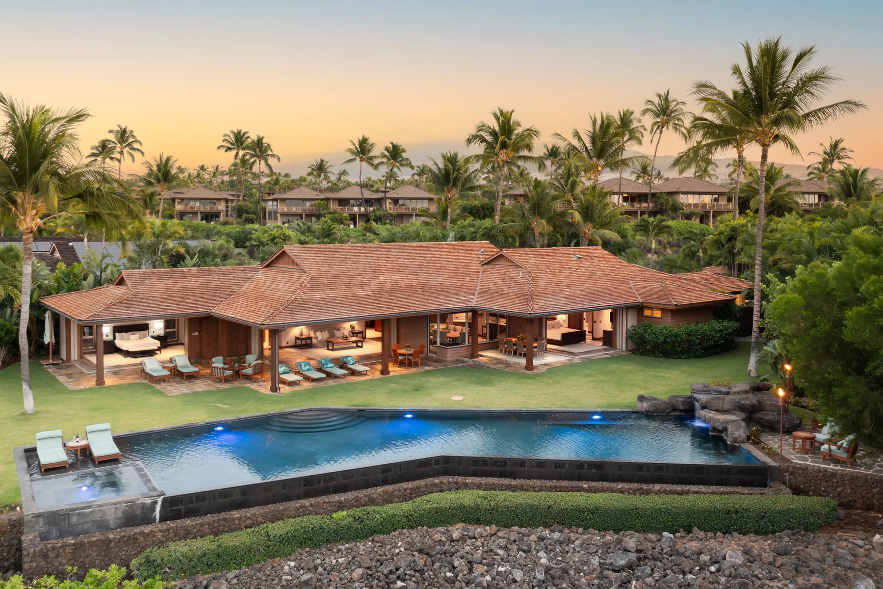 A tropical resort villa with a long swimming pool and palm trees at sunset.