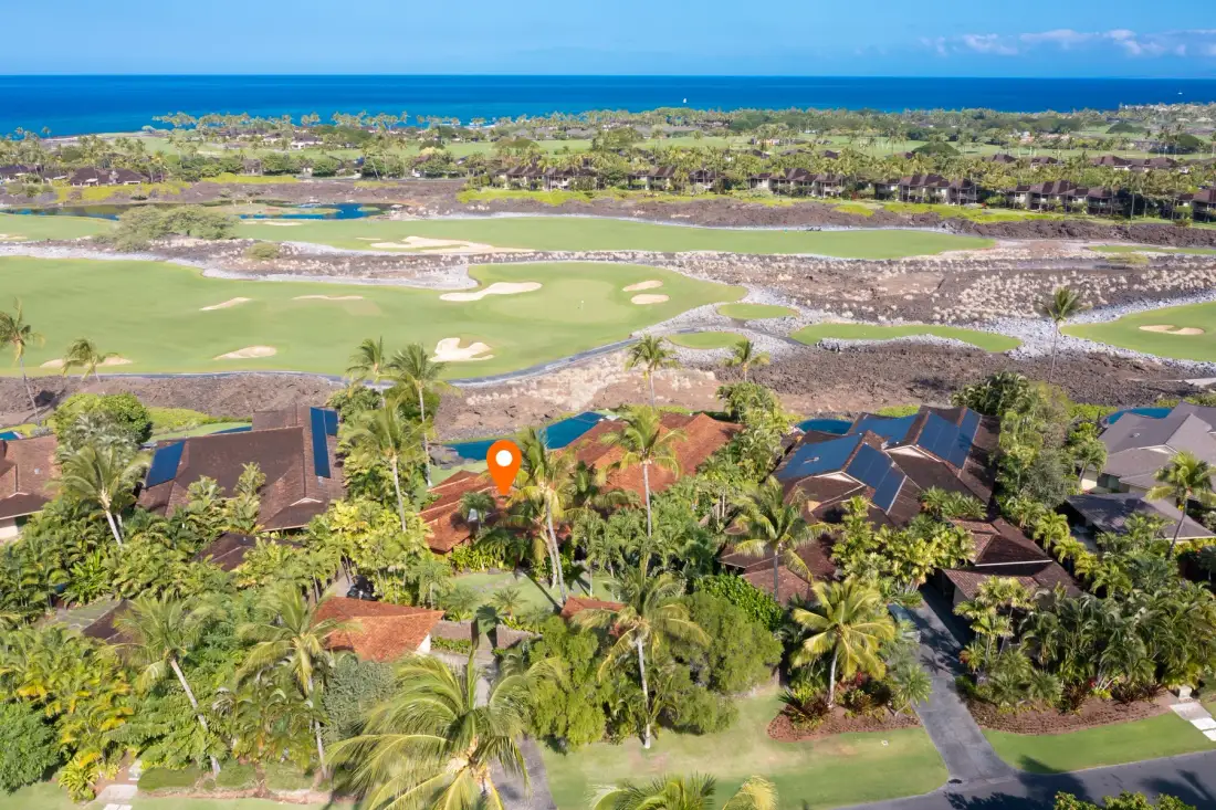 An aerial view of homes among palm trees beside a golf course, with the ocean in the distance.