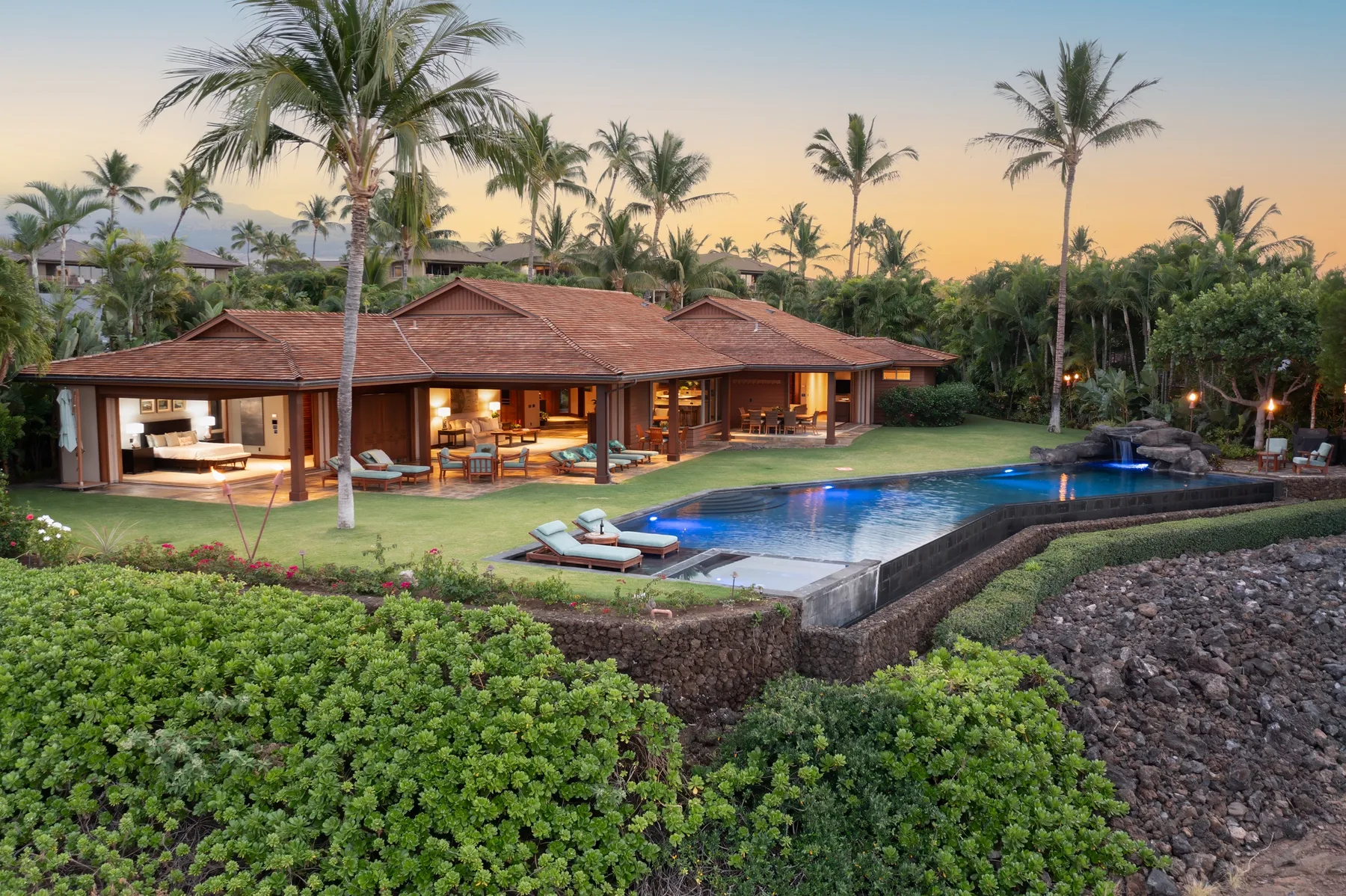 A tropical villa and swimming pool sit among palm trees and lush greenery at sunset.