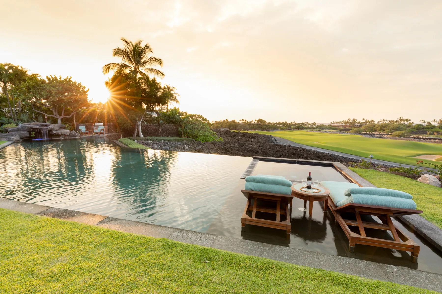 An outdoor swimming pool with cushioned loungers beside a green landscape at sunset.