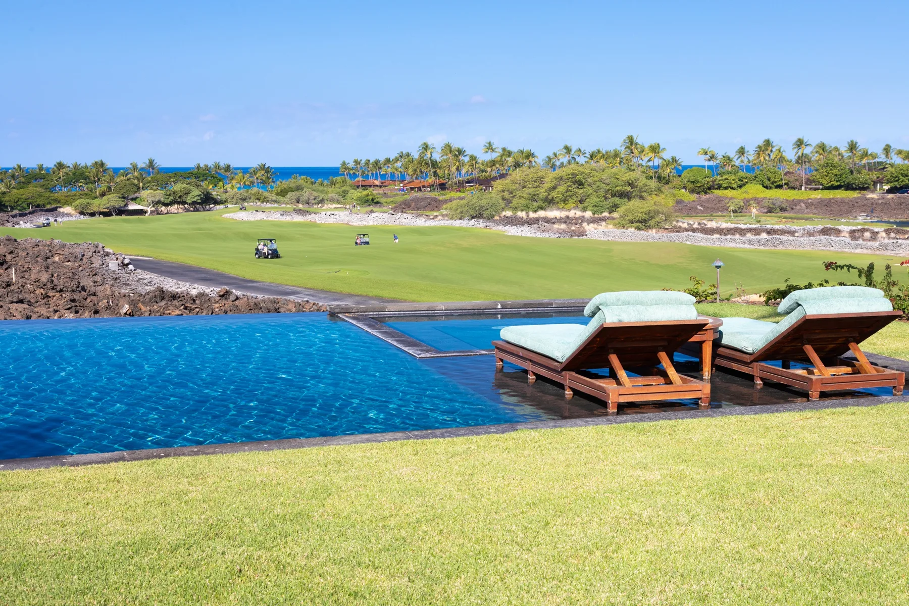 An outdoor swimming pool overlooks a grassy landscape with cushioned loungers nearby.