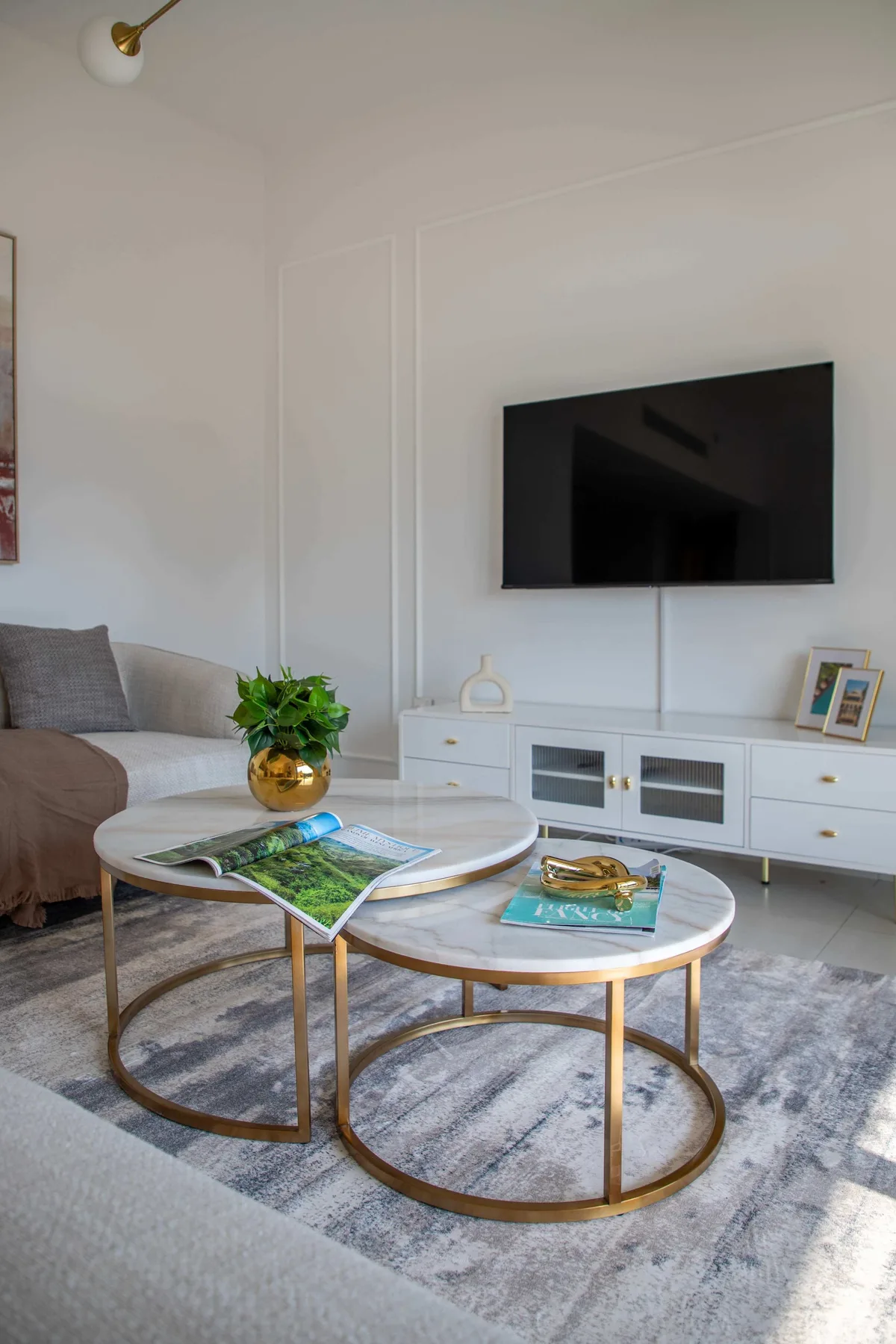 Una luminosa sala de estar tiene un televisor montado en la pared, un mueble bajo y mesas de centro redondas.