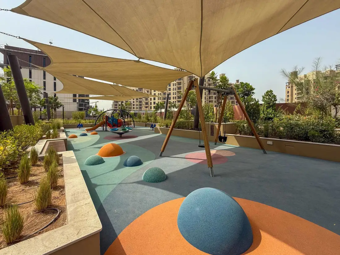 Un parque infantil al aire libre tiene suelo de colores y juegos bajo toldos.