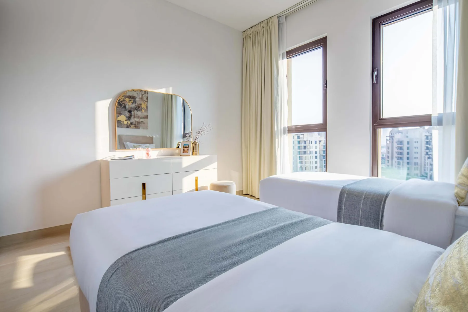 Dormitorio luminoso con dos camas junto a ventanas altas.