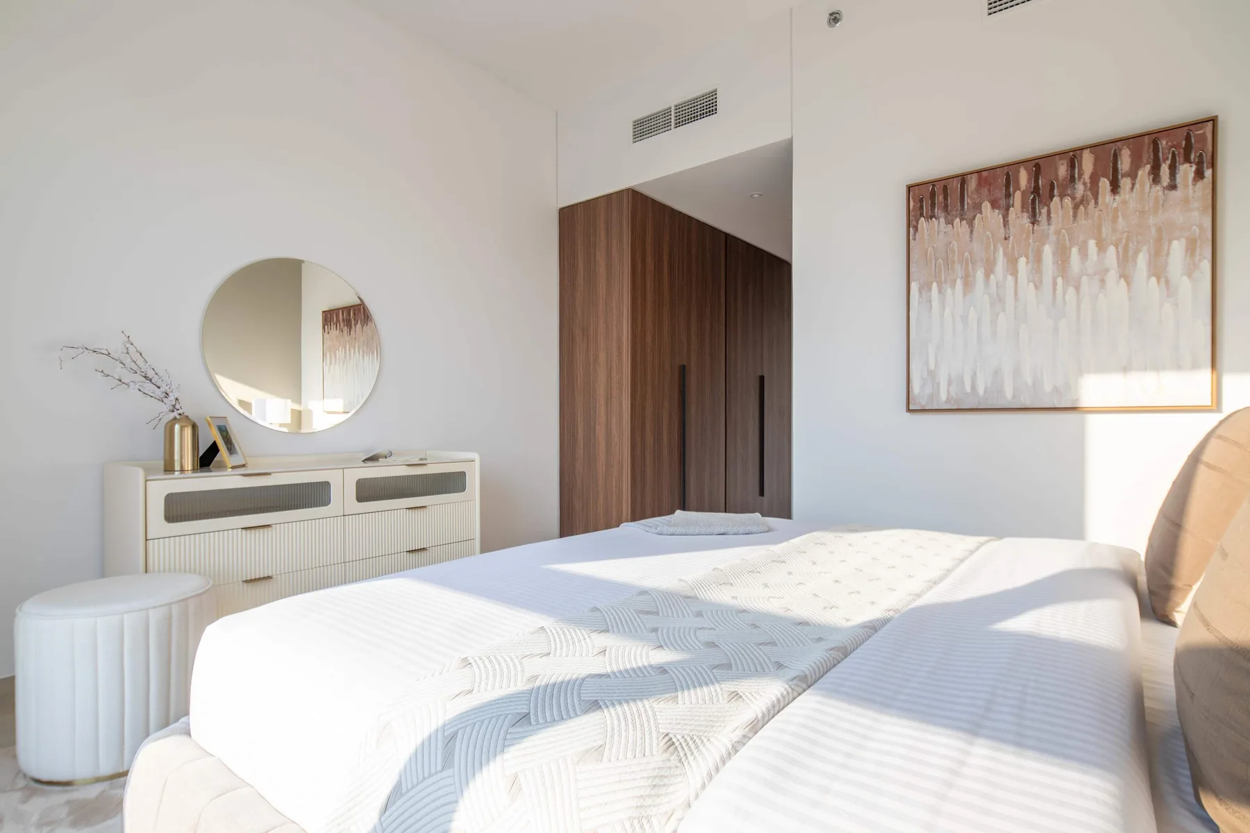 Un dormitorio luminoso con una cama tendida y un espejo redondo sobre la cómoda.