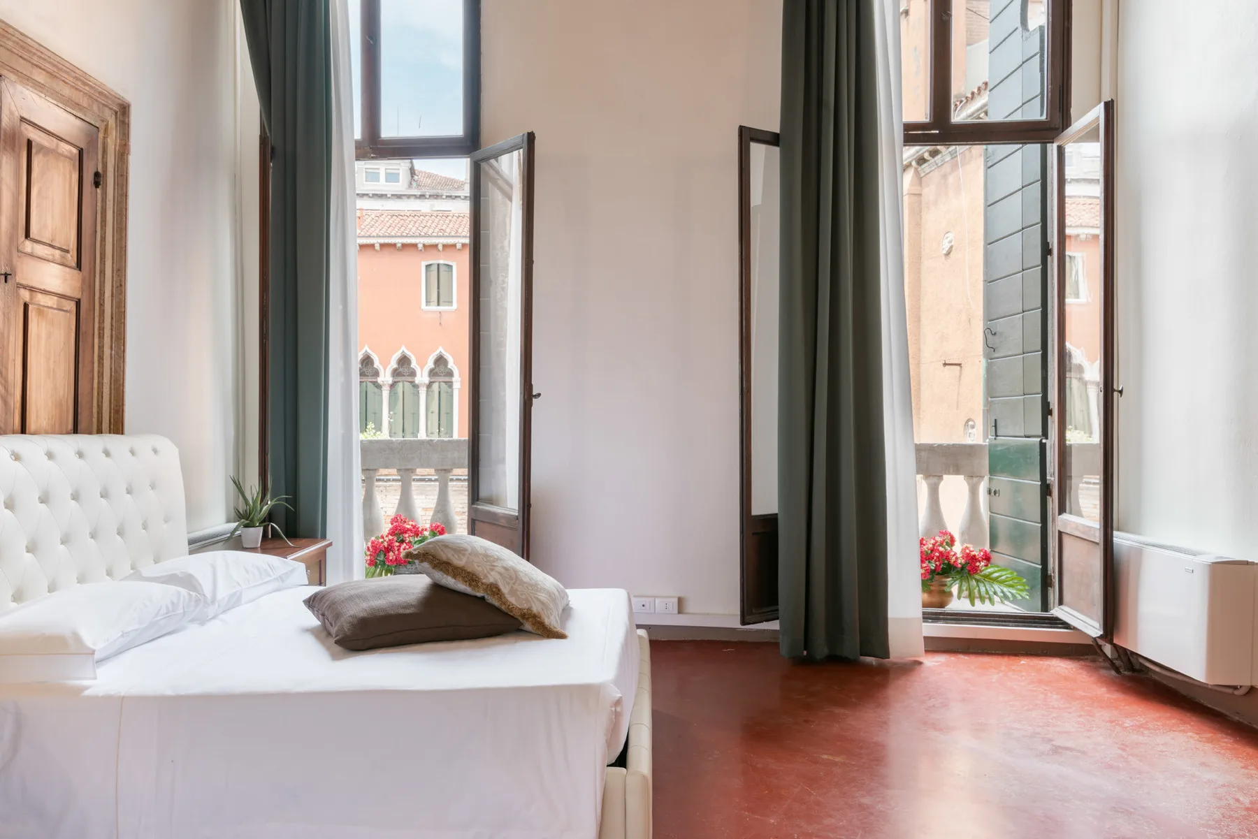 Un dormitorio con una cama junto a ventanas altas abiertas y cortinas oscuras.