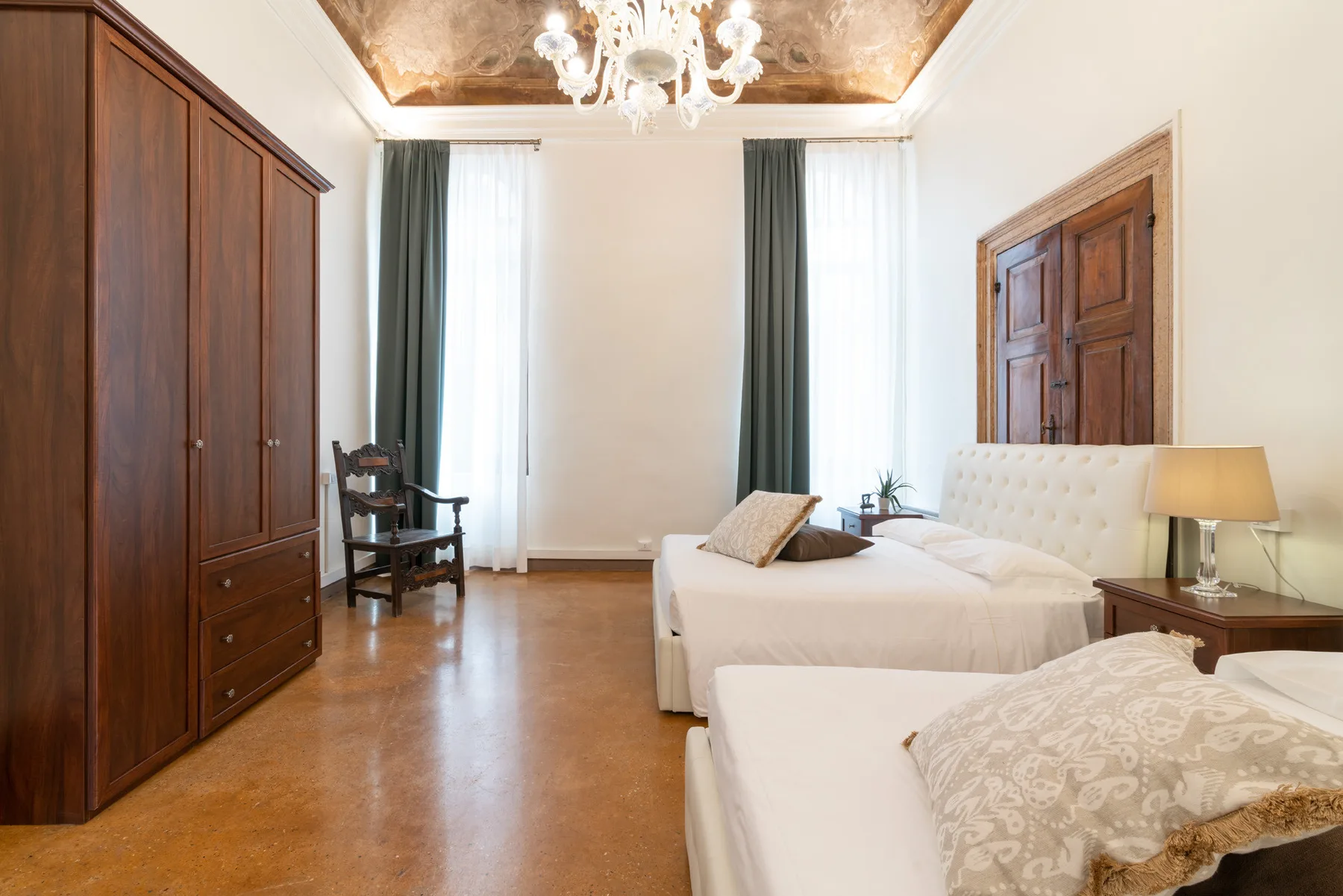 Un dormitorio luminoso con dos camas, un armario de madera y ventanas altas con cortinas.