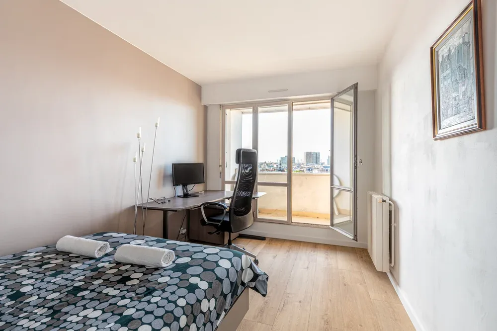 A bedroom with a bed, desk, and large windows overlooking the city.