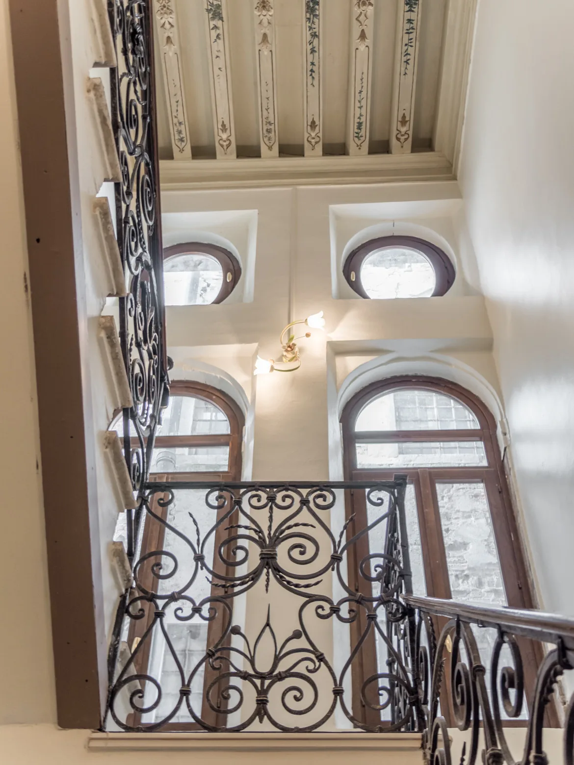 An interior stairwell with ornate iron railings and tall arched windows.