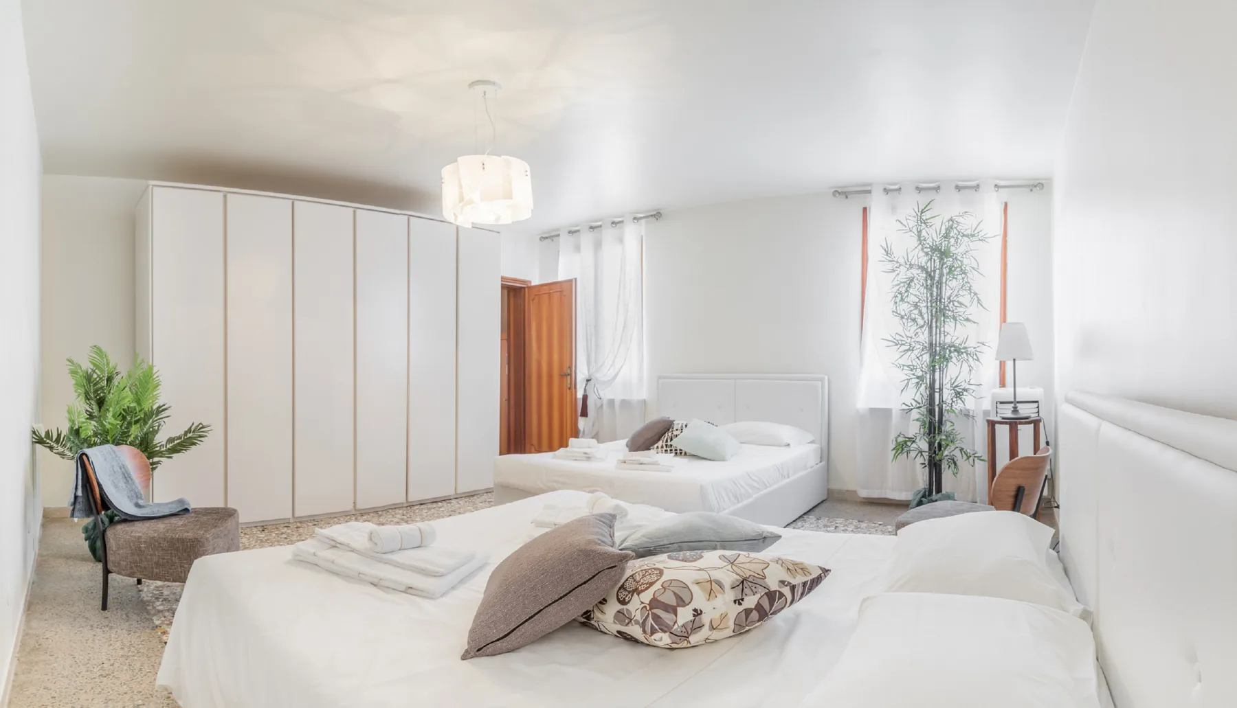 A bright bedroom with two beds, white wardrobes, and a tall plant.