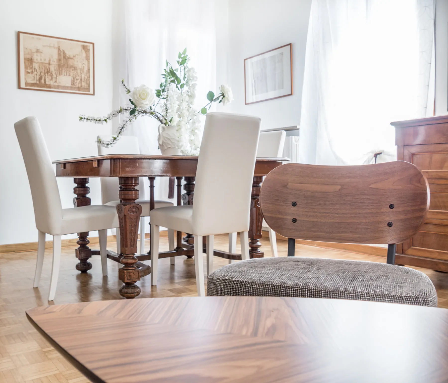 A bright dining area features a wooden table, upholstered seating, and sheer curtains.