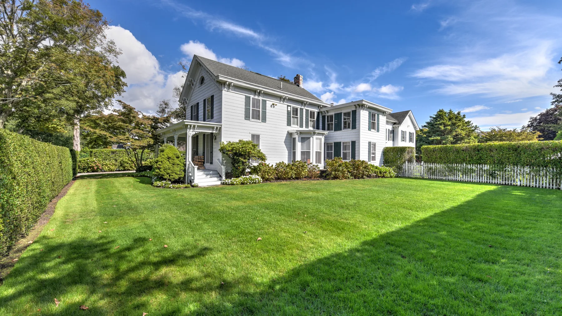 A white two-story house stands beside a broad green lawn and neatly trimmed hedges.