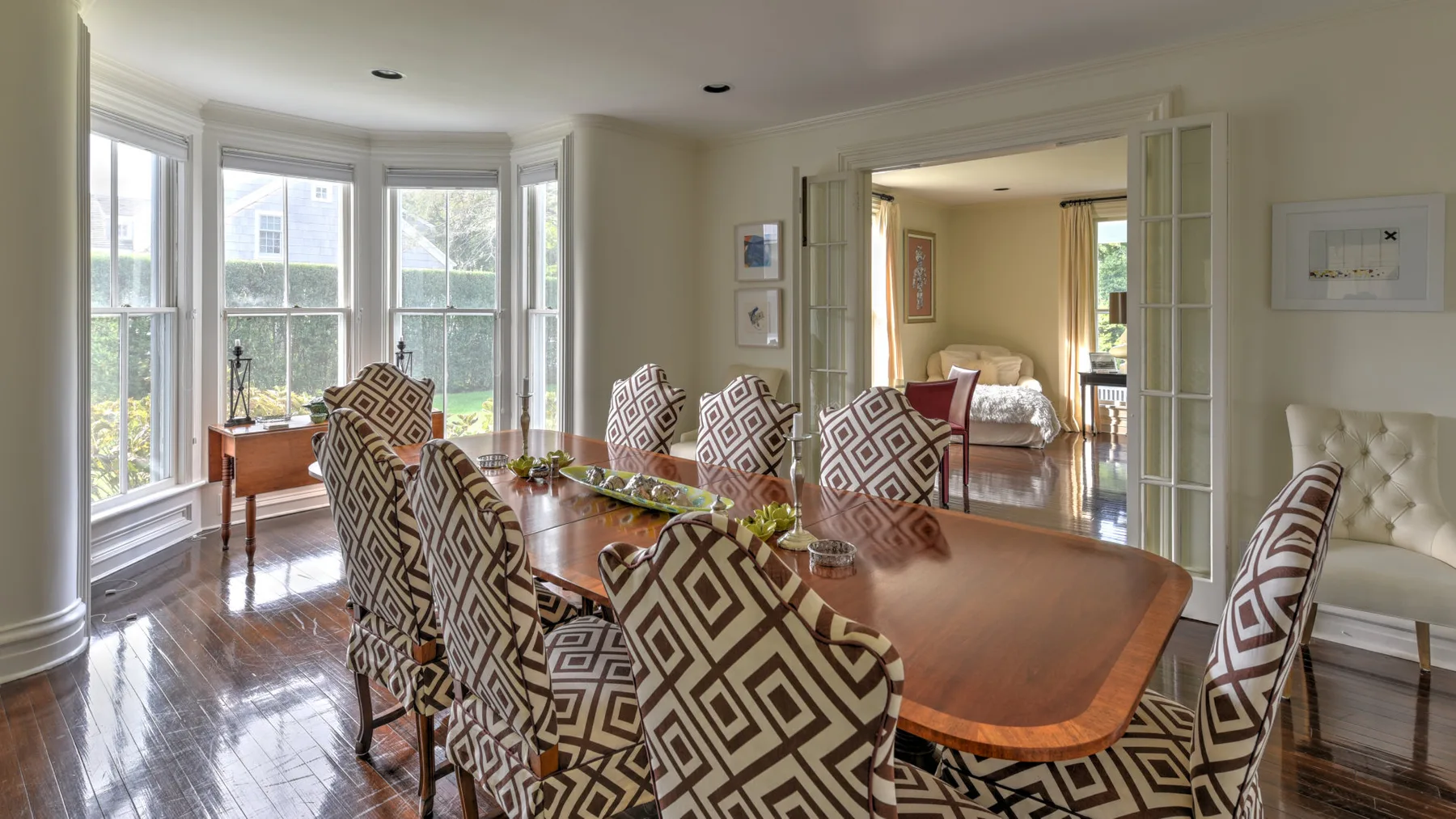 A dining room with a long wooden table and patterned seating beside bay windows.