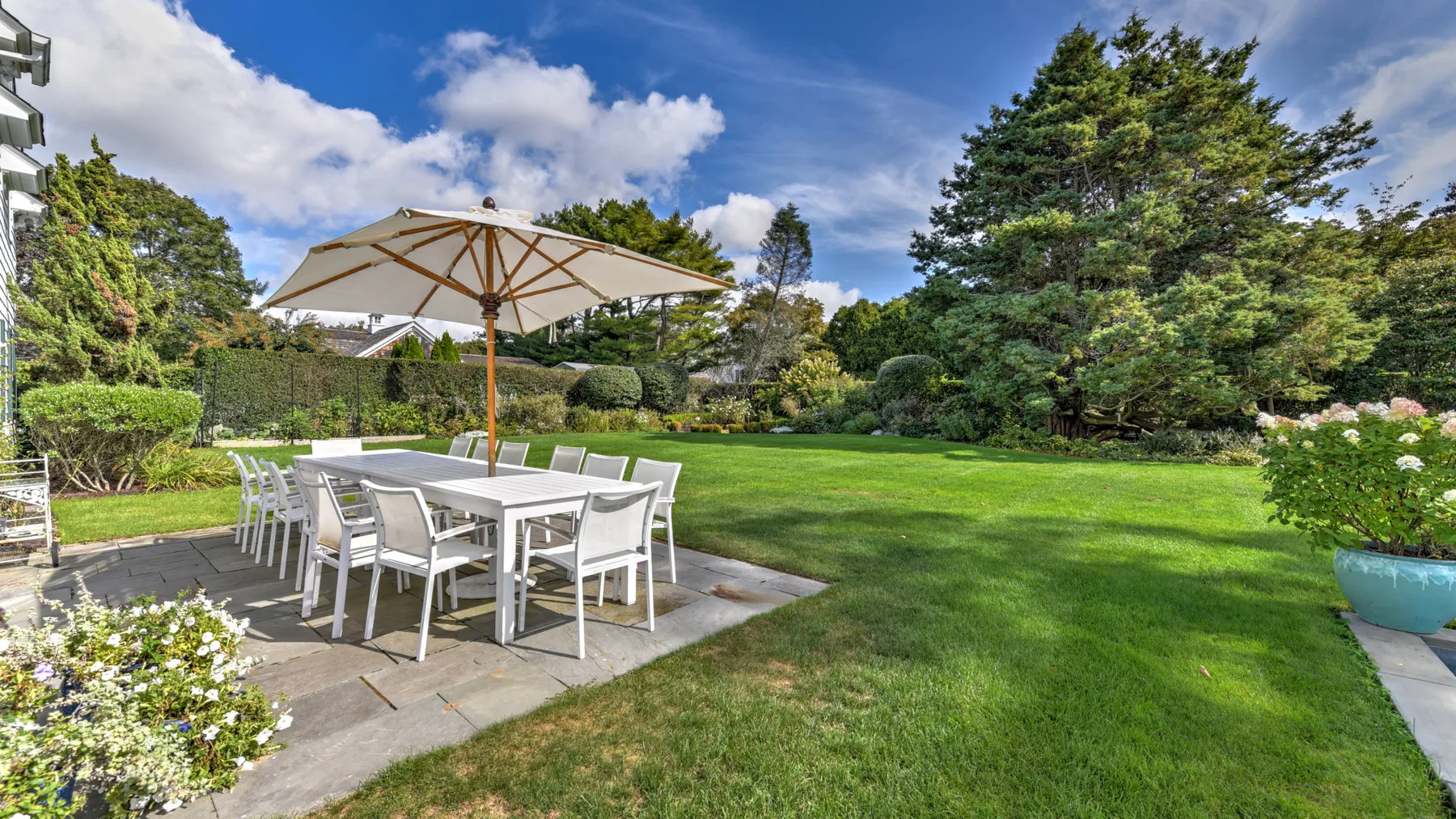 An outdoor dining area with a table beneath a large umbrella overlooks a green garden.