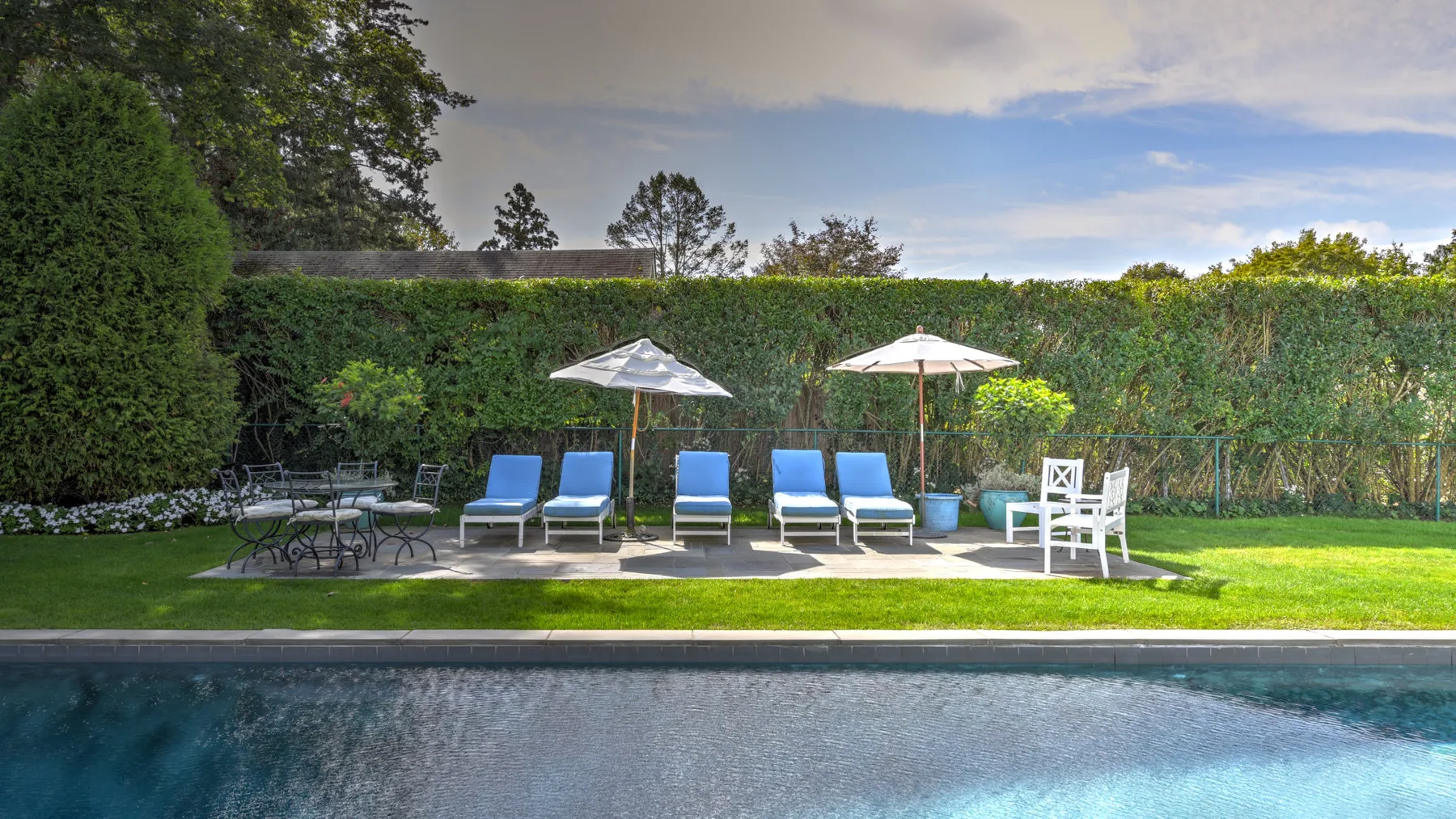 An outdoor poolside seating area with lounge chairs and umbrellas beside a hedge.