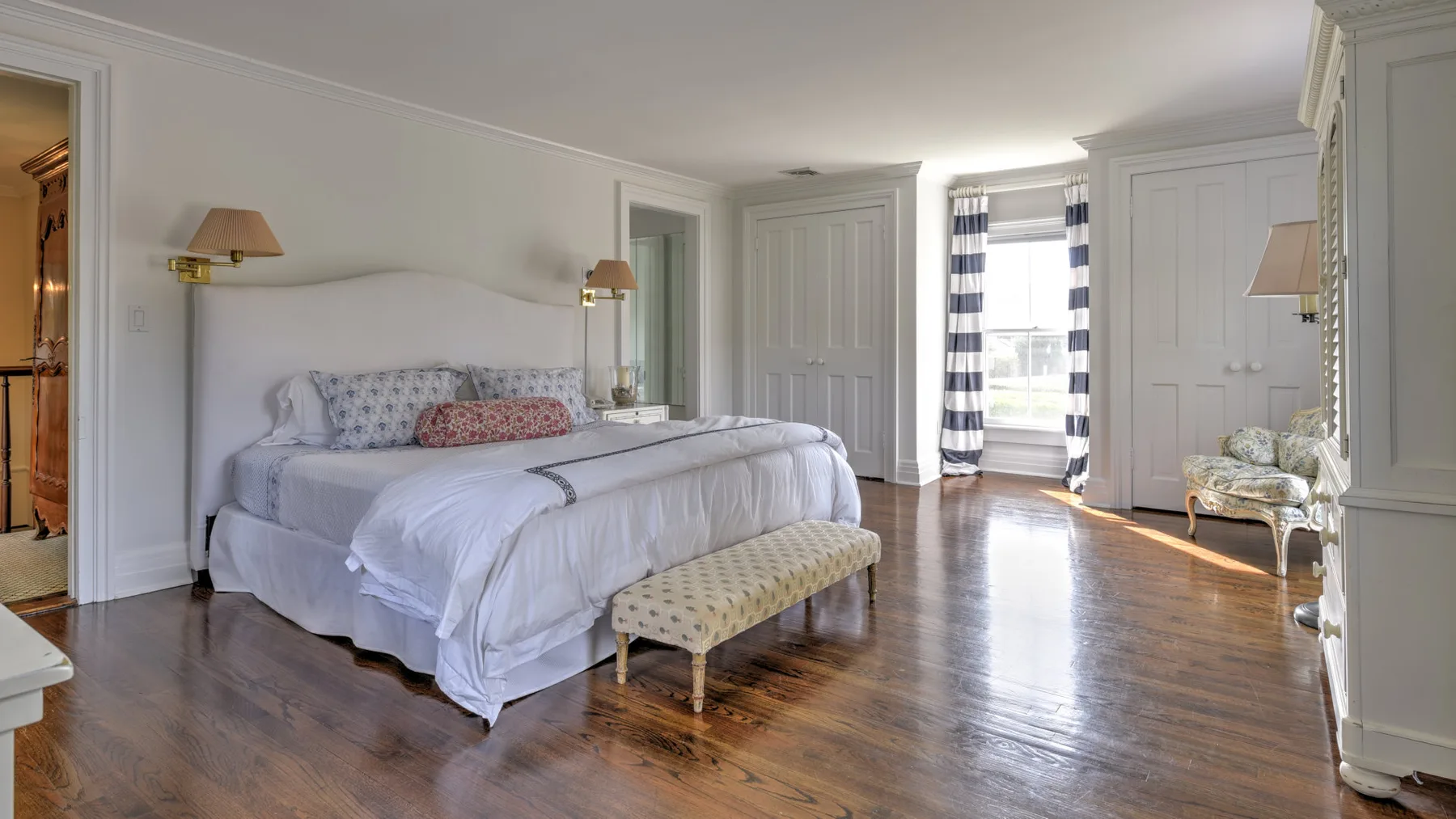 A bright bedroom with a large bed, a bench, and a window with striped curtains.