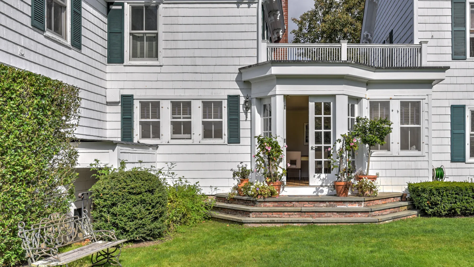 A white house opens onto a garden lawn, with a raised patio and potted plants.