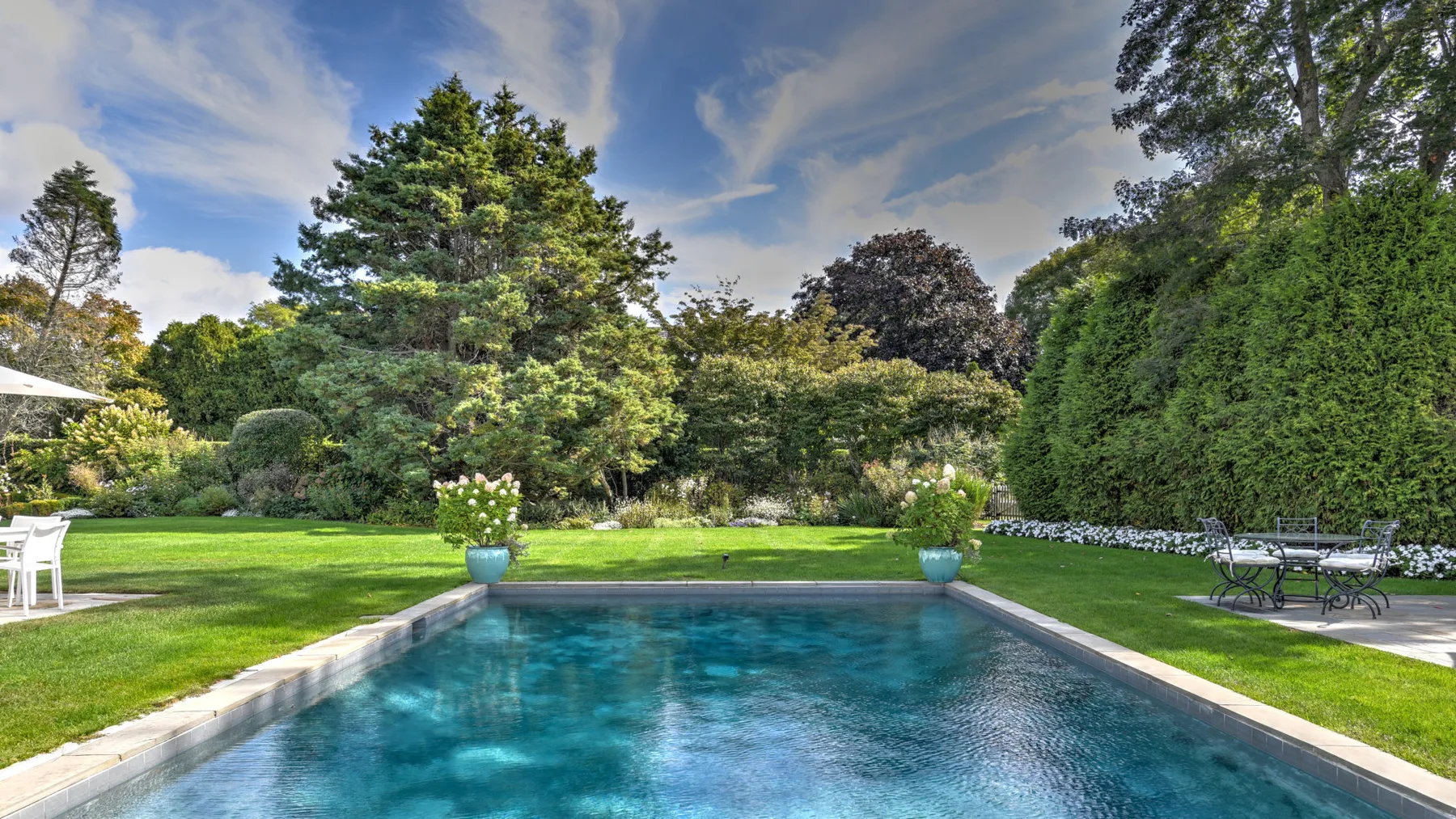 An outdoor swimming pool surrounded by a lawn, trees, and garden plants.