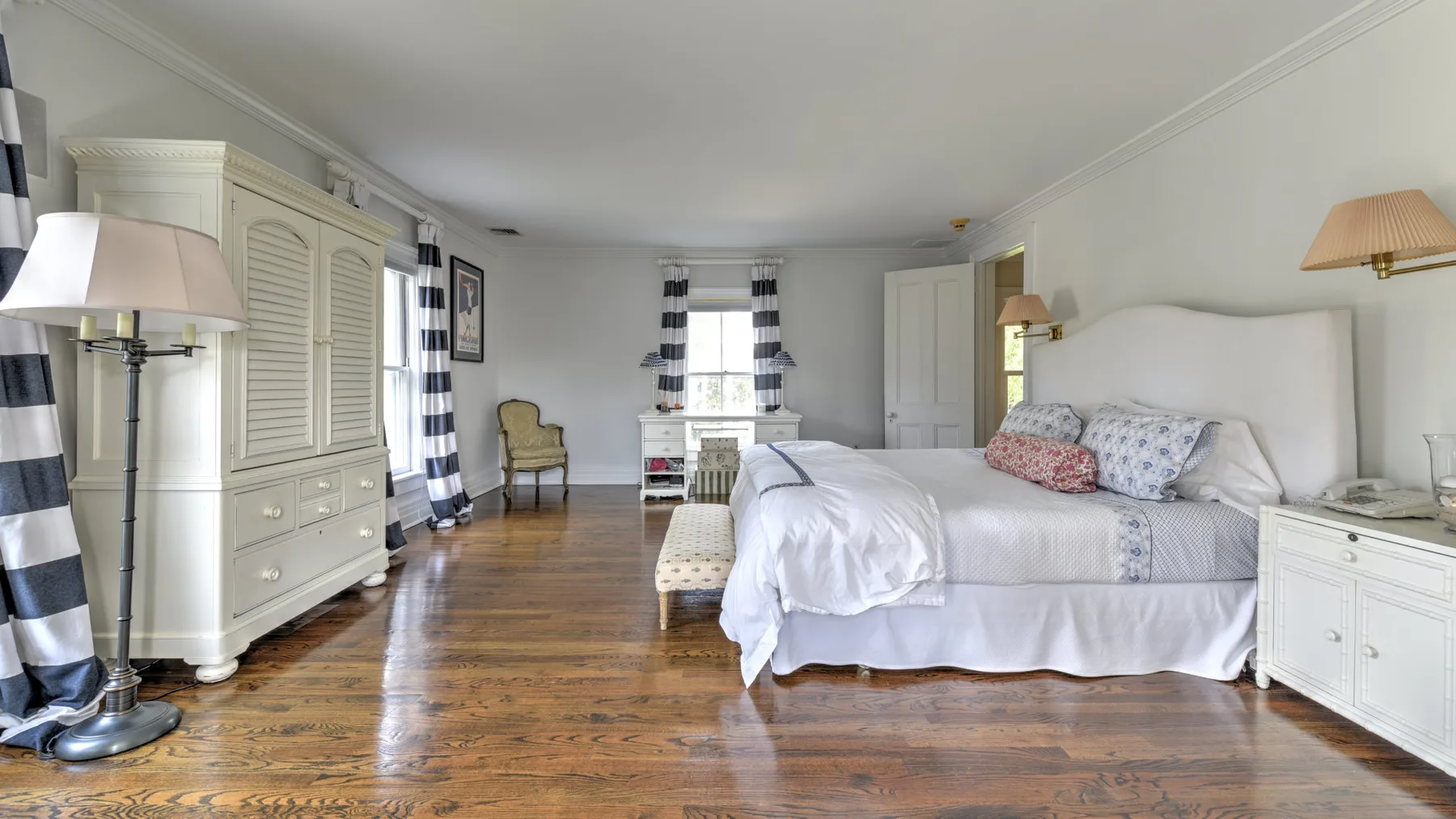 A bright bedroom with a large bed, white furnishings, and a wooden floor.