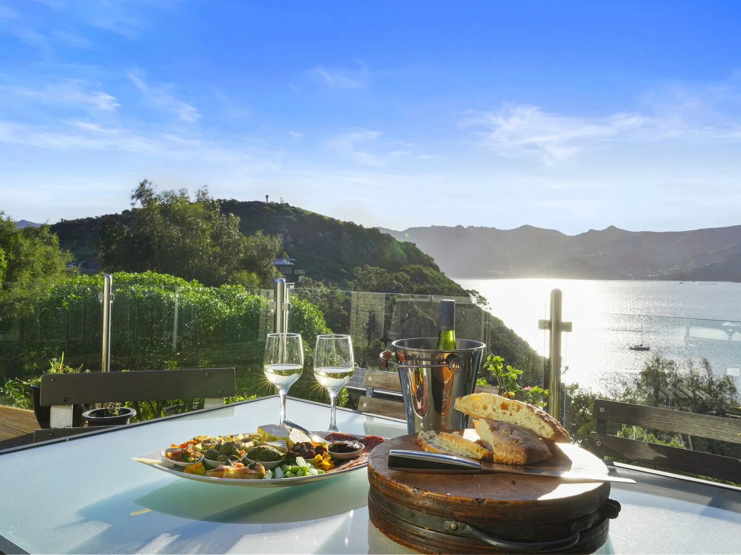 An outdoor dining table holds a meal, bread, wine glasses, and a bottle with a coastal view.