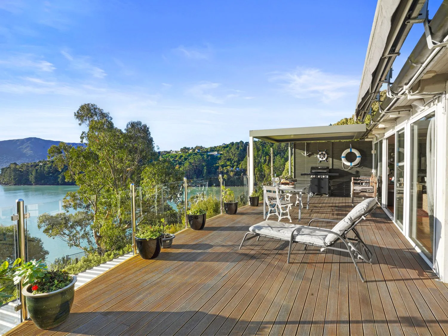An outdoor wooden deck with lounge chairs tables potted plants and a barbecue overlooking water and hills at Kaioruru View Diamond Harbour
