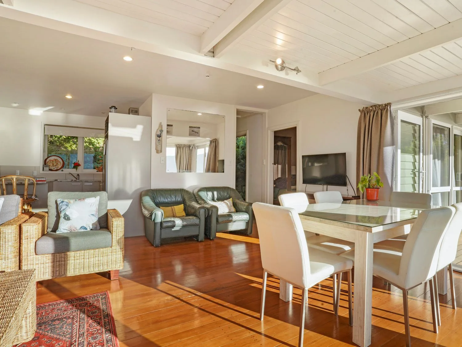 Una zona de estar y comedor con sillas de mimbre y cuero, una mesa de comedor de cristal con sillas blancas, un televisor y suelos de madera en Kaioruru View - Diamond Harbour Holiday Home, en Diamond Harbour