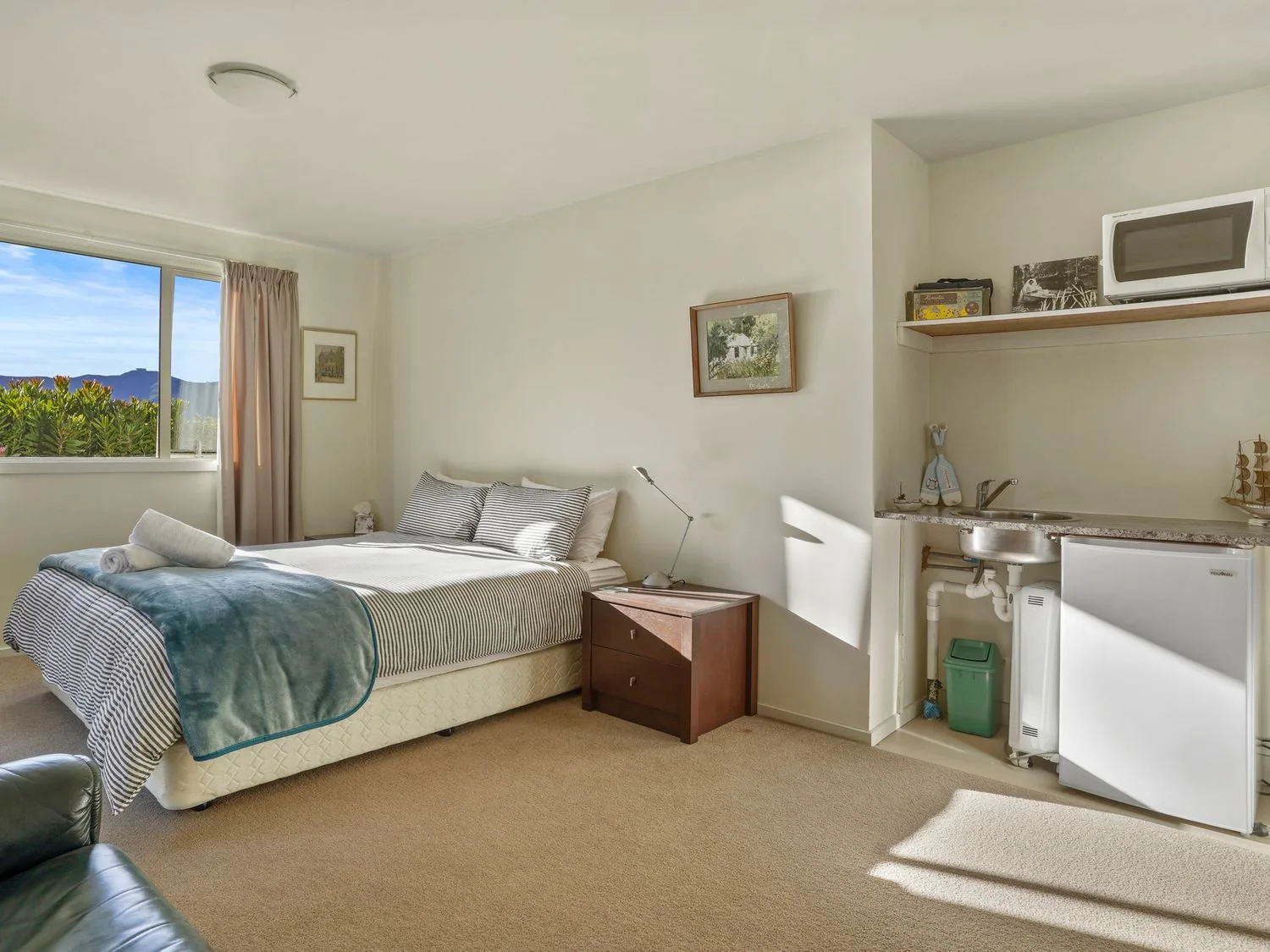 Un dormitorio con una cama cerca de una ventana, una mesita de noche y una pequeña cocina con fregadero, microondas y minirrefrigerador en Kaioruru View Diamond Harbour Holiday Home, en Diamond Harbour