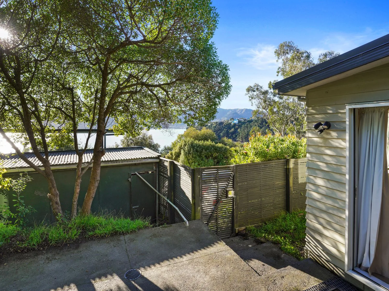 Una zona exterior con un patio de hormigón, un árbol, una valla de madera, una puerta y vistas a las colinas y al agua en Kaioruru View - Diamond Harbour Holiday Home, en Diamond Harbour